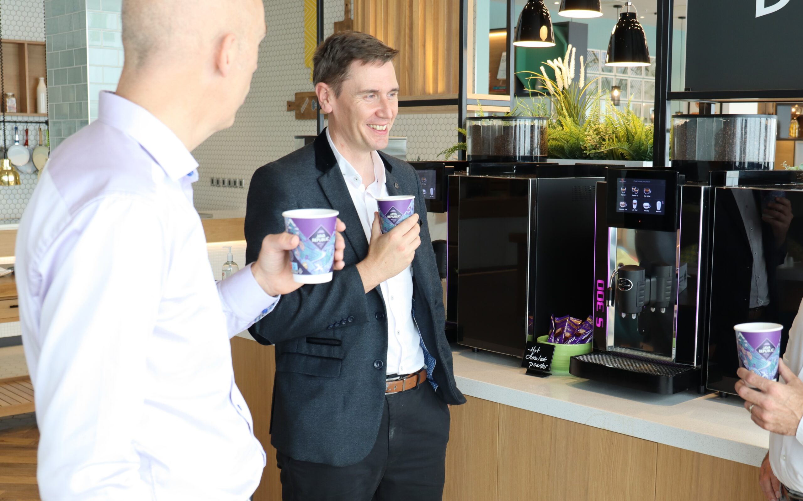 Two men holding Java Republic coffee cups smiling beside a Rex-Royal bean to cup coffee machine in an office setting